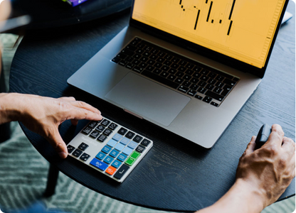 User operating the Riskeys Smart Trading Keypad next to a laptop displaying trading charts and data.